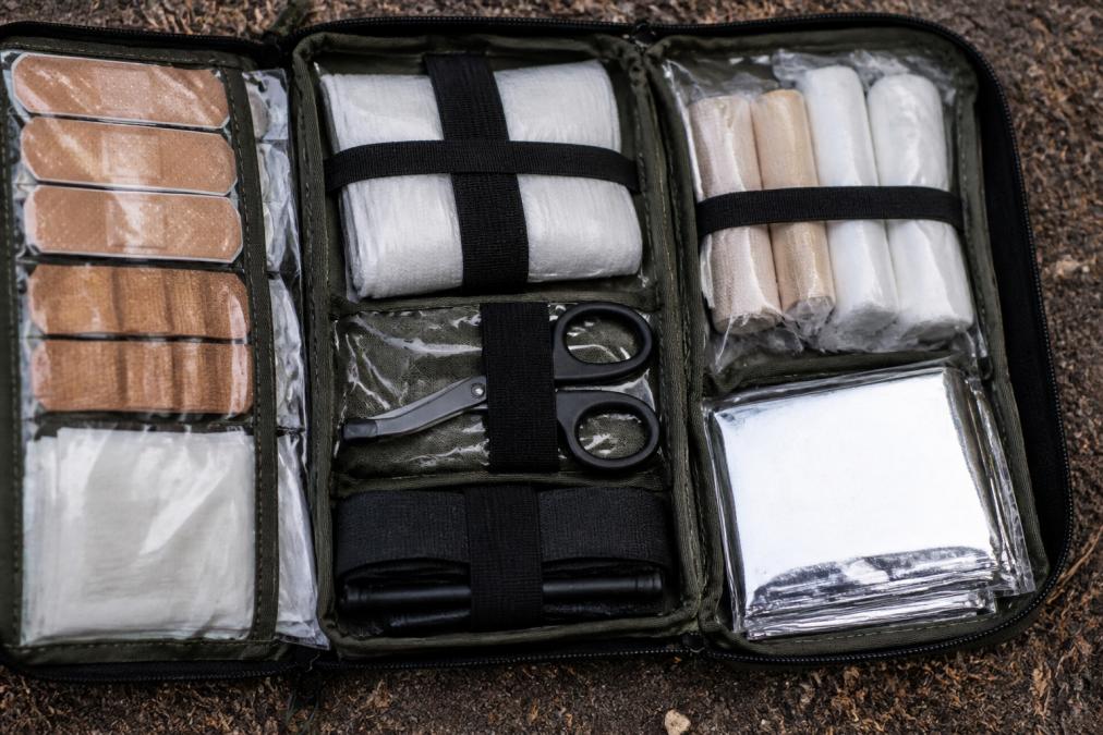 Close-up of wilderness first aid kit contents laid out systematically, showing adhesive bandages, sterile gauze pads, trauma shears, tourniquets, and emergency blanket organized in waterproof compartments