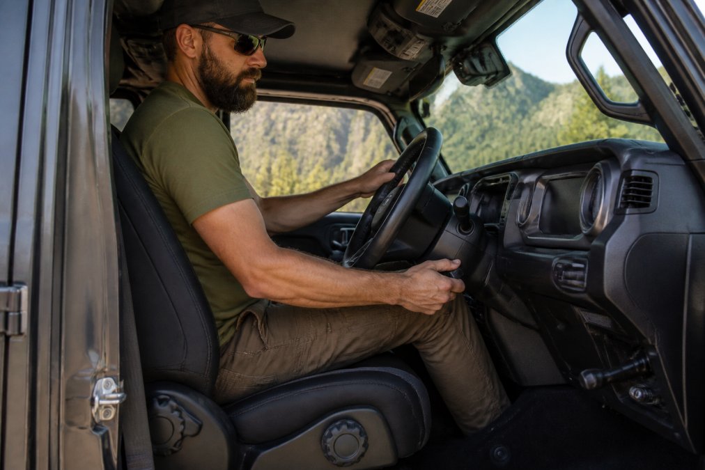 A tall male driver seated in a rugged off-road SUV interior, adjusting the steering column with one hand, showing a clear view of the cockpit layout and seat position, natural daylight coming through the windshield