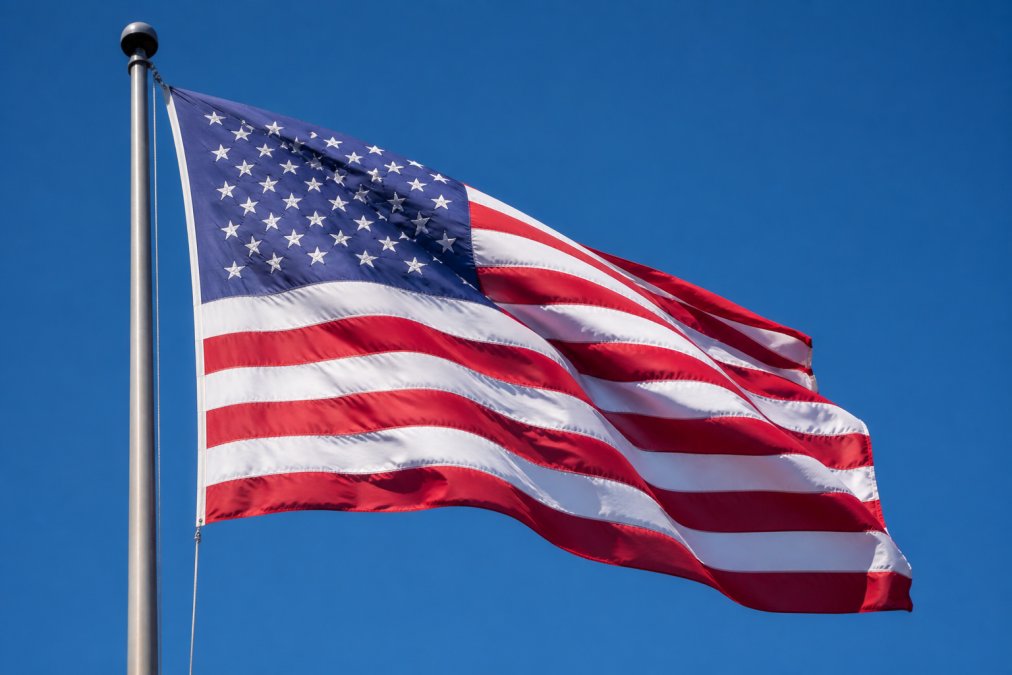 Close-up of a large American flag made from heavy-duty polyester fabric flying vigorously on a tall flagpole against a clear blue sky, fabric fully extended and rippling in strong wind