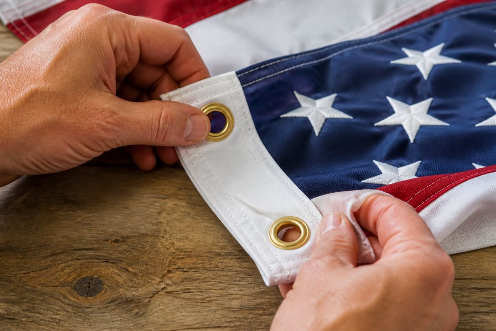 Hands inspecting the reinforced stitched edge and brass grommets of an American flag laid flat on a wooden table, close detail showing the canvas header and double-fold hem stitching in natural daylight