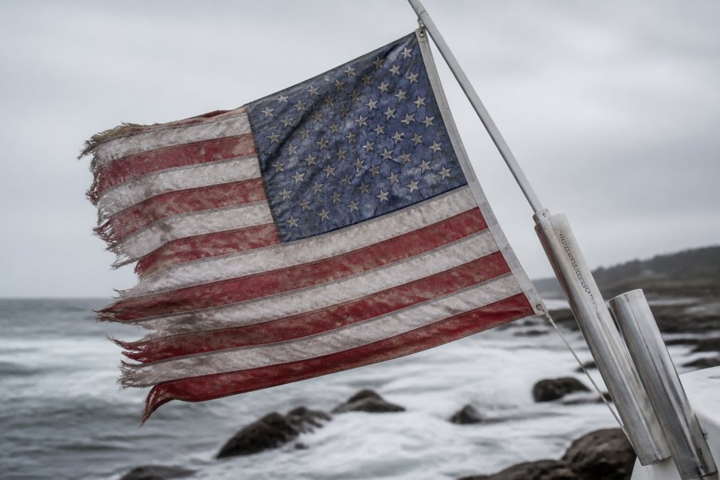 Close-up of a weathered, salt-stained flag with frayed edges and faded colors hanging on a coastal flagpole with ocean spray visible and waves crashing in the background