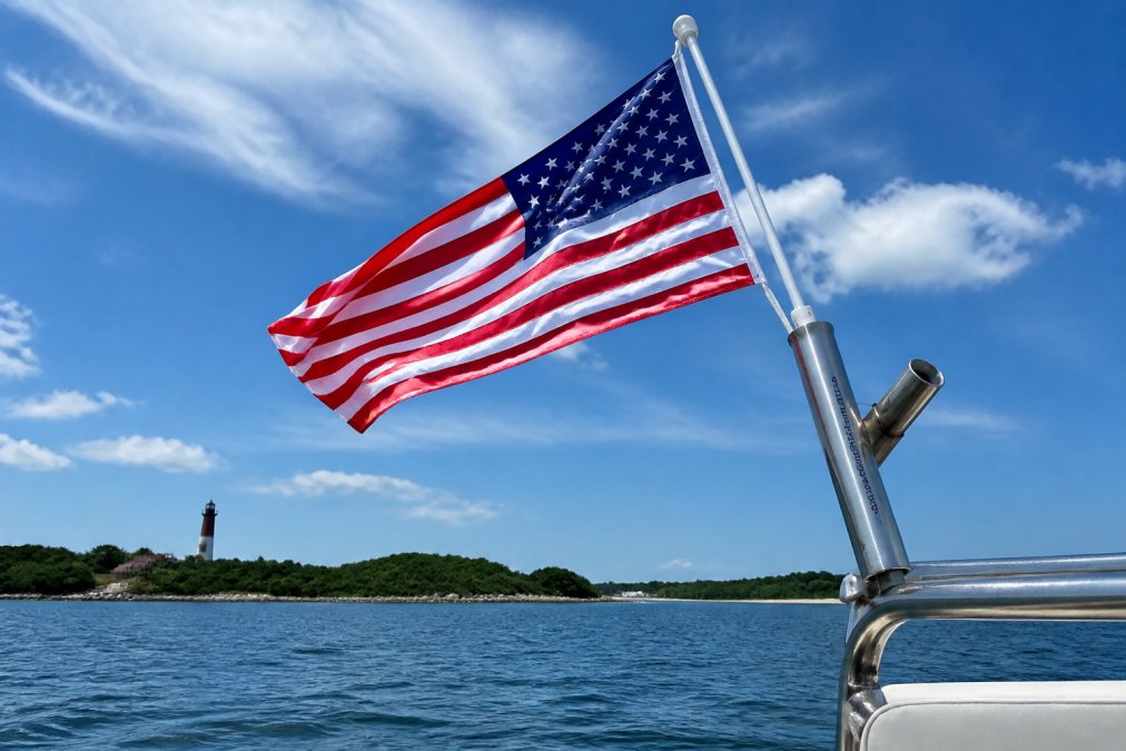 A vibrant, well-maintained nylon flag with rich colors flying proudly on a marine-grade stainless steel flagpole against a bright blue sky with coastal scenery and a lighthouse visible in the background