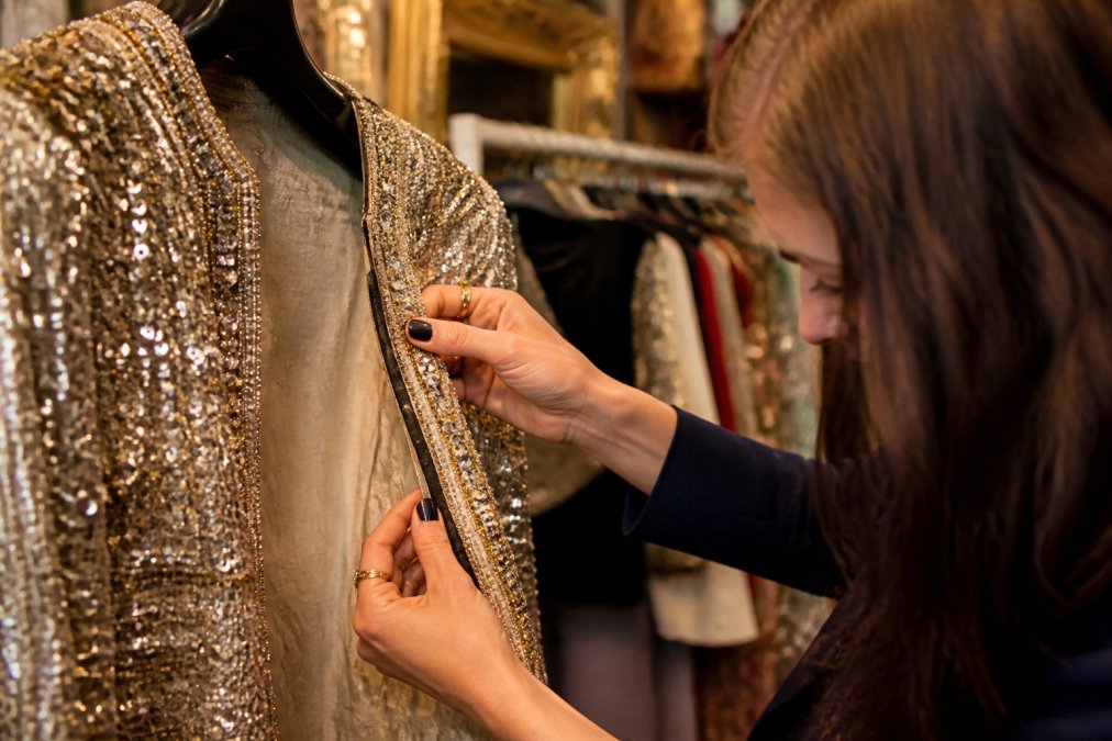Close-up of a woman's hands examining the beaded trim and interior seam of a vintage sequined jacket under warm boutique lighting