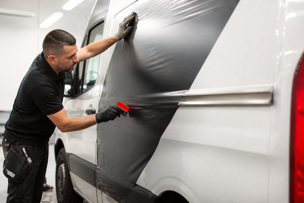 Professional technician carefully applying vinyl wrap to commercial fleet vehicle, showing precision technique and attention to detail during installation with proper lighting and clean workspace