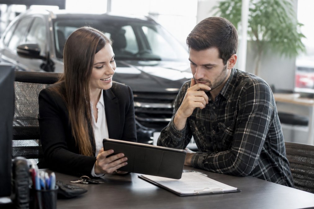A friendly auto finance specialist sitting with a customer at a dealership desk, reviewing flexible payment plan options on a tablet, warm and reassuring office environment