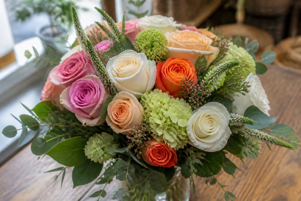 Close-up of vibrant fresh-cut floral arrangement with roses, greenery, and complementary blooms displayed on a wooden table with soft natural lighting