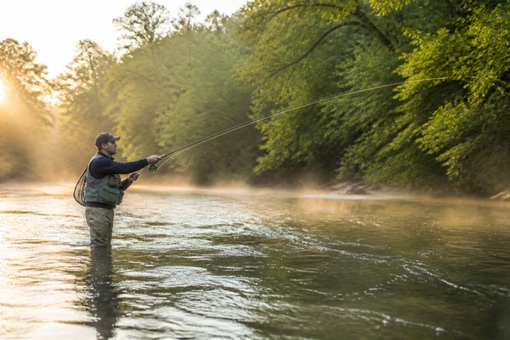 A fly angler standing knee-deep in a calm North Carolina river at sunrise, casting a fly rod with mist rising off the water and trees lining the bank