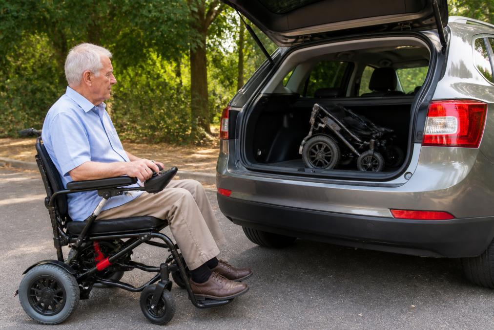 Older adult in a lightweight folding electric wheelchair positioned next to a car boot, demonstrating how easily the chair fits for travel and transport, with natural daylight showing the compact folded size