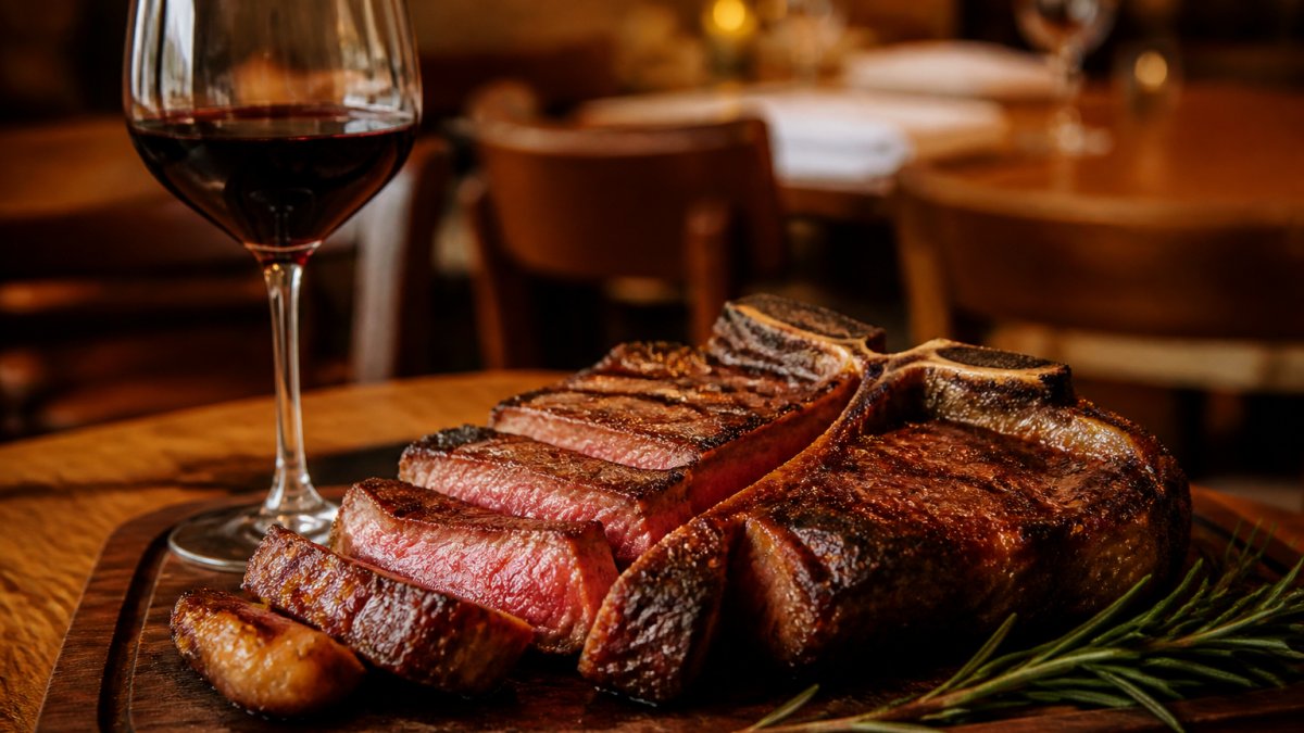A thick grilled Florentine T-bone steak resting on a wooden board, sliced to reveal a pink centre, with a glass of red wine and fresh rosemary beside it under warm restaurant lighting
