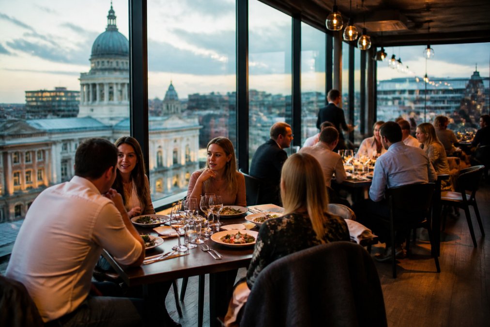 Modern restaurant dining room with floor-to-ceiling windows overlooking the city centre, warm ambient lighting reflecting off polished wooden tables, and diners engaged in conversation over plated courses