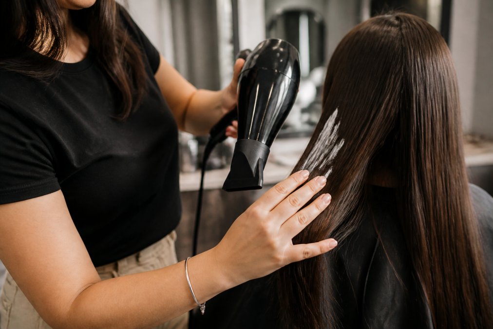 Close-up of someone using a blow dryer and smoothing cream on freshly treated hair, demonstrating proper styling technique with fingers guiding hair through the heat