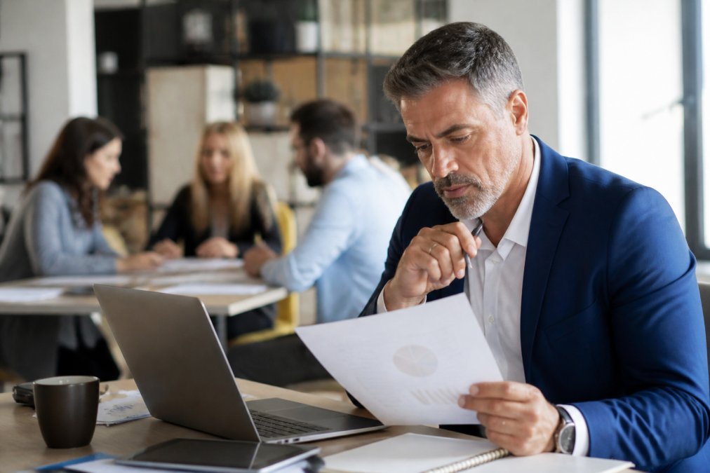 Business professional reviewing marketing strategy documents and analytics on a laptop with team members collaborating in background in modern office