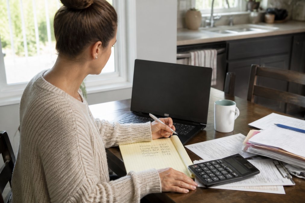 A homeowner sitting at a kitchen table with a laptop open to a cleaning cost calculator, holding a pen and notepad with room measurements written down, natural light from a window