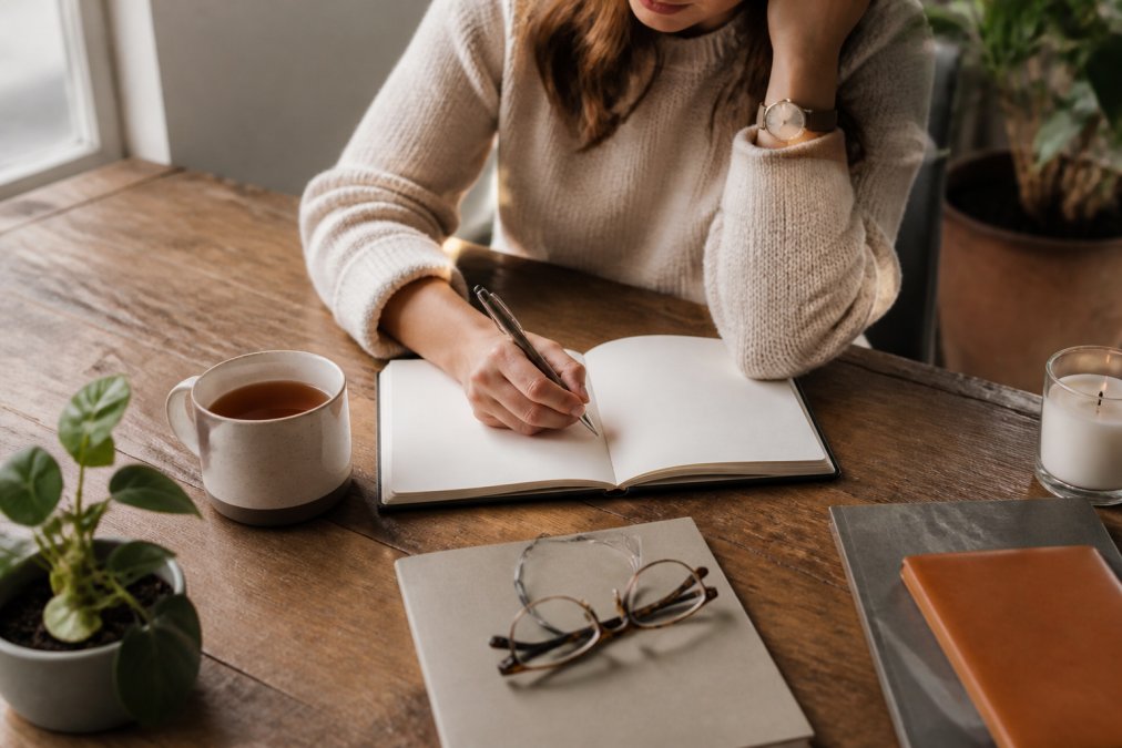 Person writing in a journal at a wooden desk with a warm cup of tea nearby, pen in hand, focused and calm, soft natural light from a nearby window illuminating the page