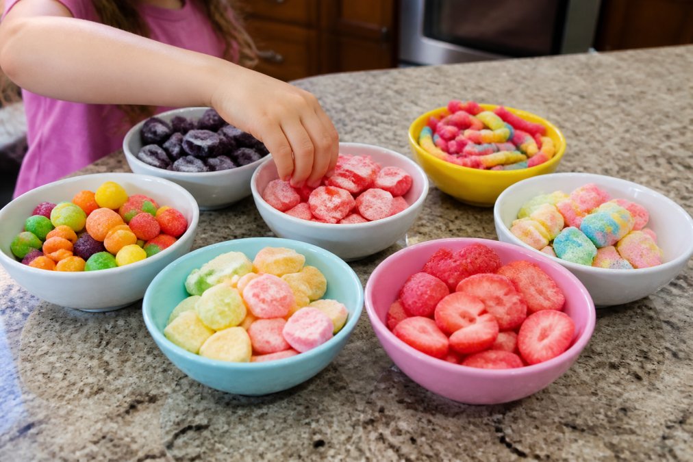 A bright, colorful flat-lay photo of several small bowls filled with different freeze dried candies in vibrant colors, with a child's hand reaching for a piece, set on a cheerful kitchen counter
