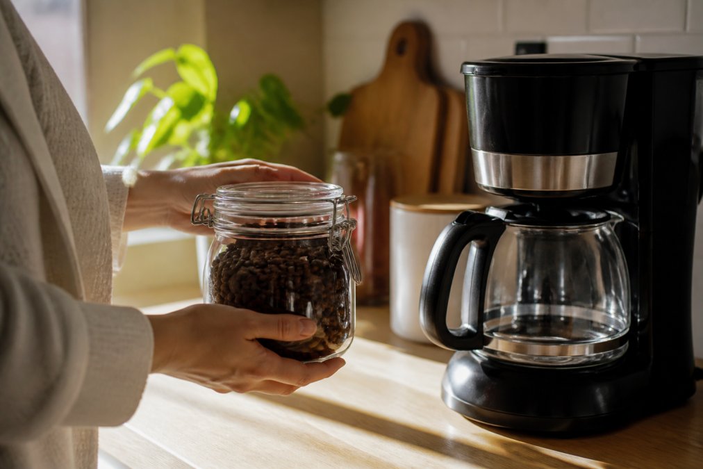Hands holding an airtight glass container filled with whole coffee beans, positioned next to a drip coffee maker on a light wood kitchen counter with morning sunlight illuminating the scene
