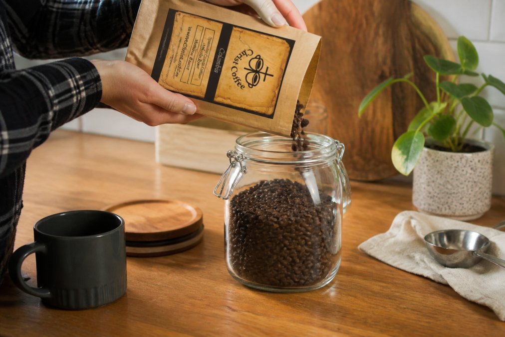 Hands placing whole coffee beans into an airtight glass container on a kitchen counter, with a roast date label visible on the container