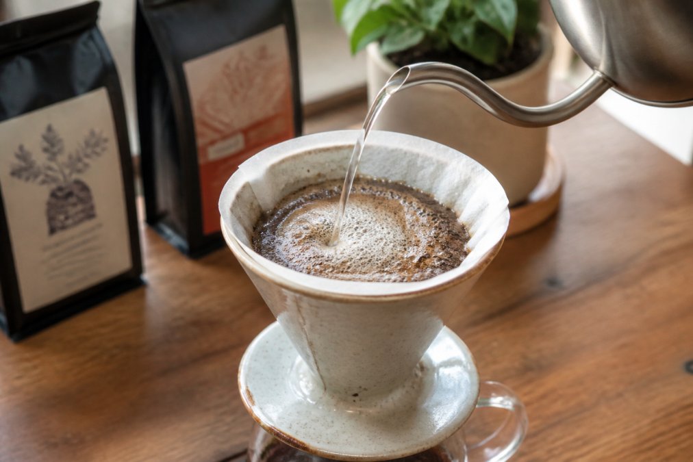 Close-up of a ceramic pour over dripper on a glass carafe, with freshly ground coffee blooming as hot water is poured in a slow circular motion, warm natural light, two Cenia's Coffee bags visible softly out of focus in the background