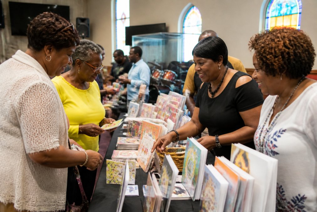 Church members gathered around a table displaying colorful greeting cards, stationery, and gift items for sale, with volunteers smiling and helping customers select products from African American Expressions collection