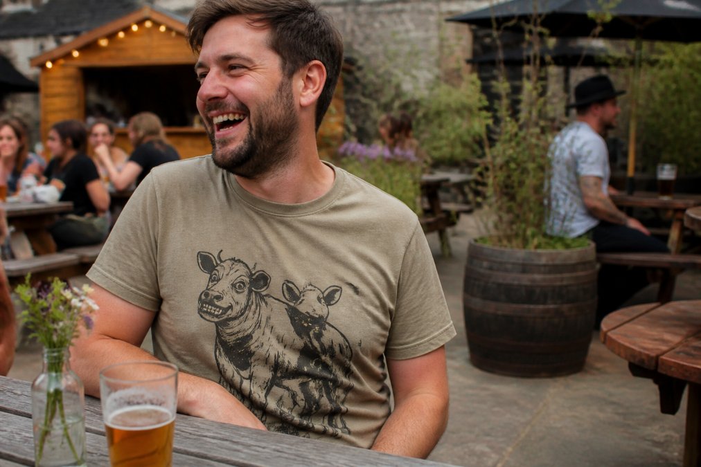 A man in his 30s wearing a graphic t-shirt with a bold animal print, laughing in a casual pub garden setting. The focus is on the t-shirt design and its fit.