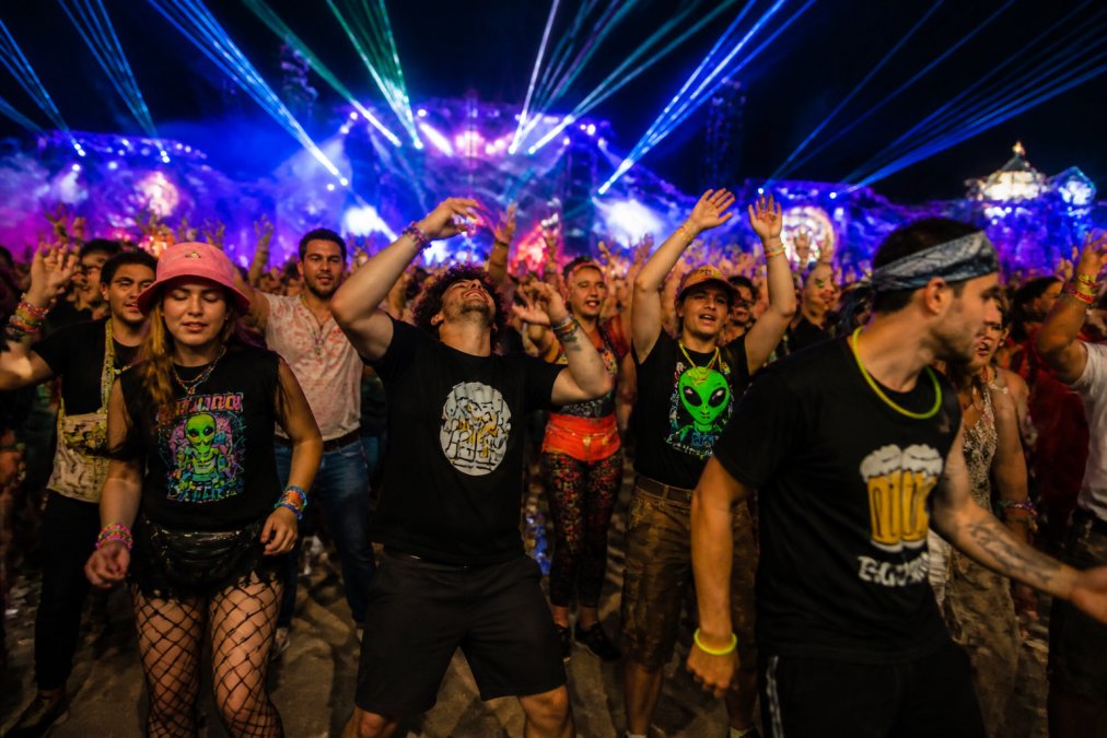 Crowd of people dancing energetically at a nighttime electronic music festival under bright neon lights and laser beams, wearing bold graphic tees and glow sticks, with dynamic movement and vibrant colors