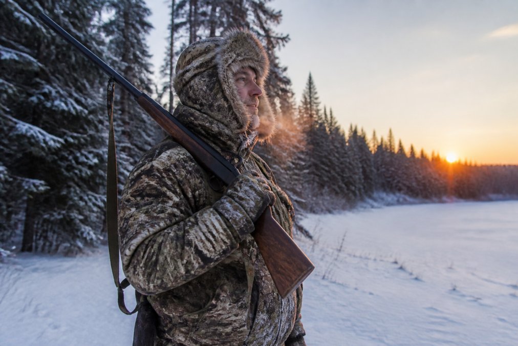 A hunter in full camouflage wearing a coyote fur trapper hat with earflaps secured under chin, standing at the edge of a snow-covered forest clearing at dawn, breath visible in cold air, rifle rested over shoulder