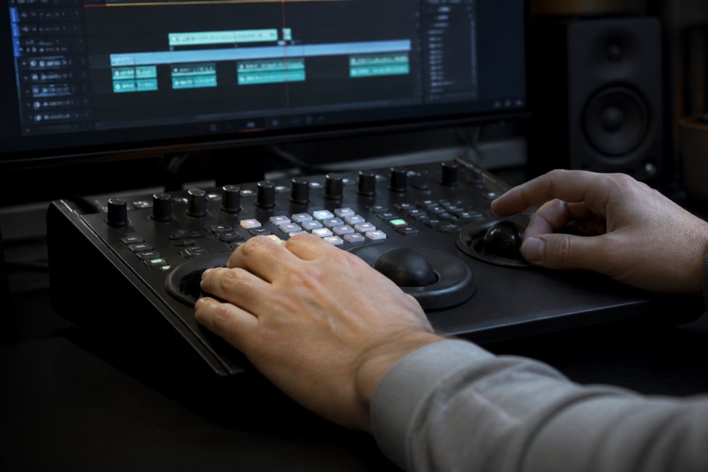 Close-up of hands using an editing console with illuminated LCD buttons and tactile knobs, video editing timeline visible on a monitor in soft studio lighting