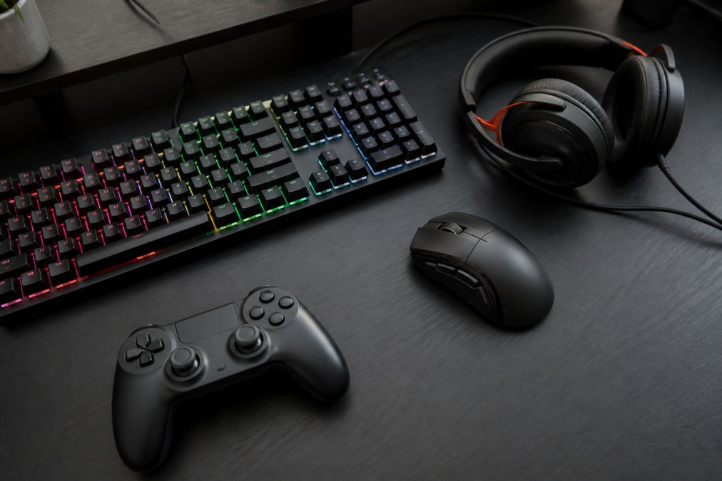 Flat lay arrangement of mechanical keyboard with RGB backlighting, precision gaming mouse with side buttons, wired gaming headset, and wireless controller on a dark gaming desk surface with cable management