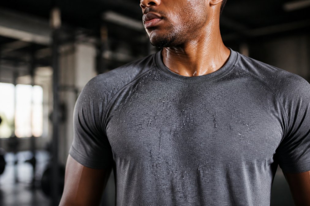 Close-up of an athlete mid-workout wearing a moisture-wicking performance tee, sweat visibly beading on the fabric surface, gym lighting catching the texture of the knit