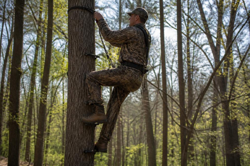 Hunter in camouflage ascending a tree using climbing sticks with saddle platform attached, tether rope secured and harness properly positioned, showing controlled ascent in natural woodland lighting