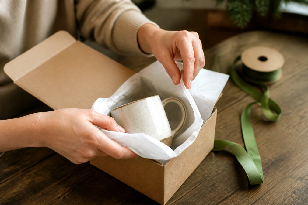 Hands carefully wrapping a ceramic mug in white tissue paper, with a kraft gift box open on a wooden surface and a ribbon nearby, showing professional gift presentation technique