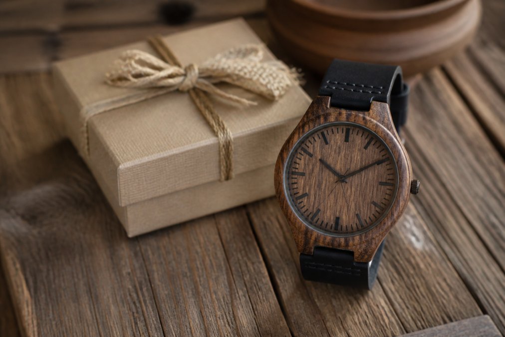 A close-up of a wooden watch with rich sandalwood grain and a black leather strap resting on a rustic wooden table beside a small gift box