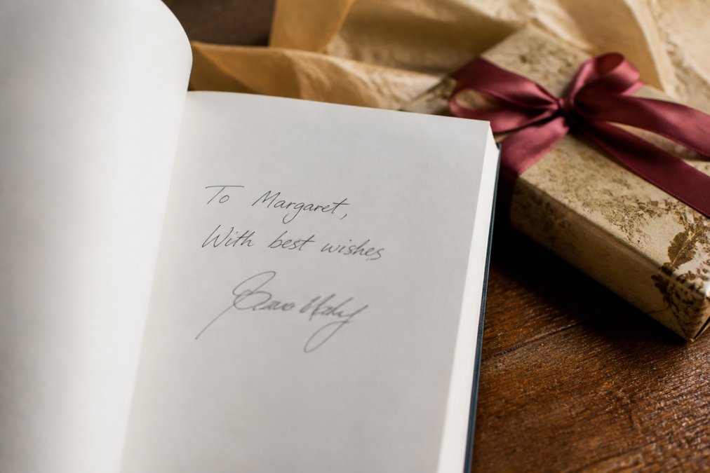 Close-up of an author-signed book dedication page with elegant handwriting, soft focus background showing wrapped gift with ribbon and tissue paper
