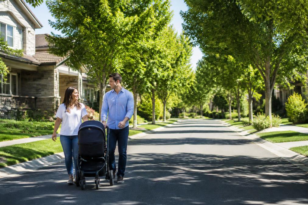 Young family walking together on a tree-lined residential street with green lawns, mature trees, and modern homes in the background, bright morning sunlight