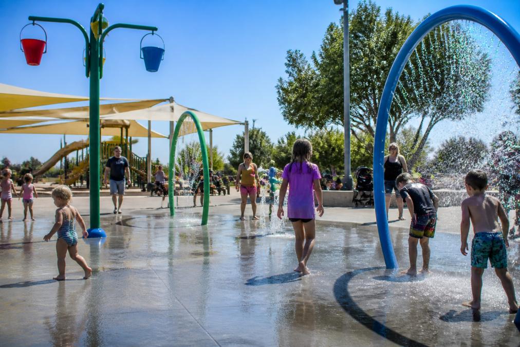 Children playing at a community park with splash pads, playground equipment, and families enjoying outdoor recreation on a sunny afternoon