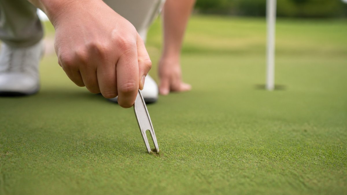 Close-up of a golfer's hands using a divot repair tool to lift and level a ball mark on a manicured putting green, with the flagstick visible in the soft-focus background