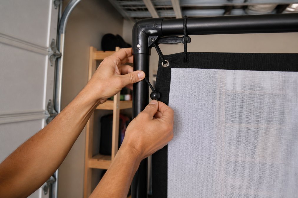 Hands attaching a golf simulator enclosure impact screen to an aluminum frame with bungee cords