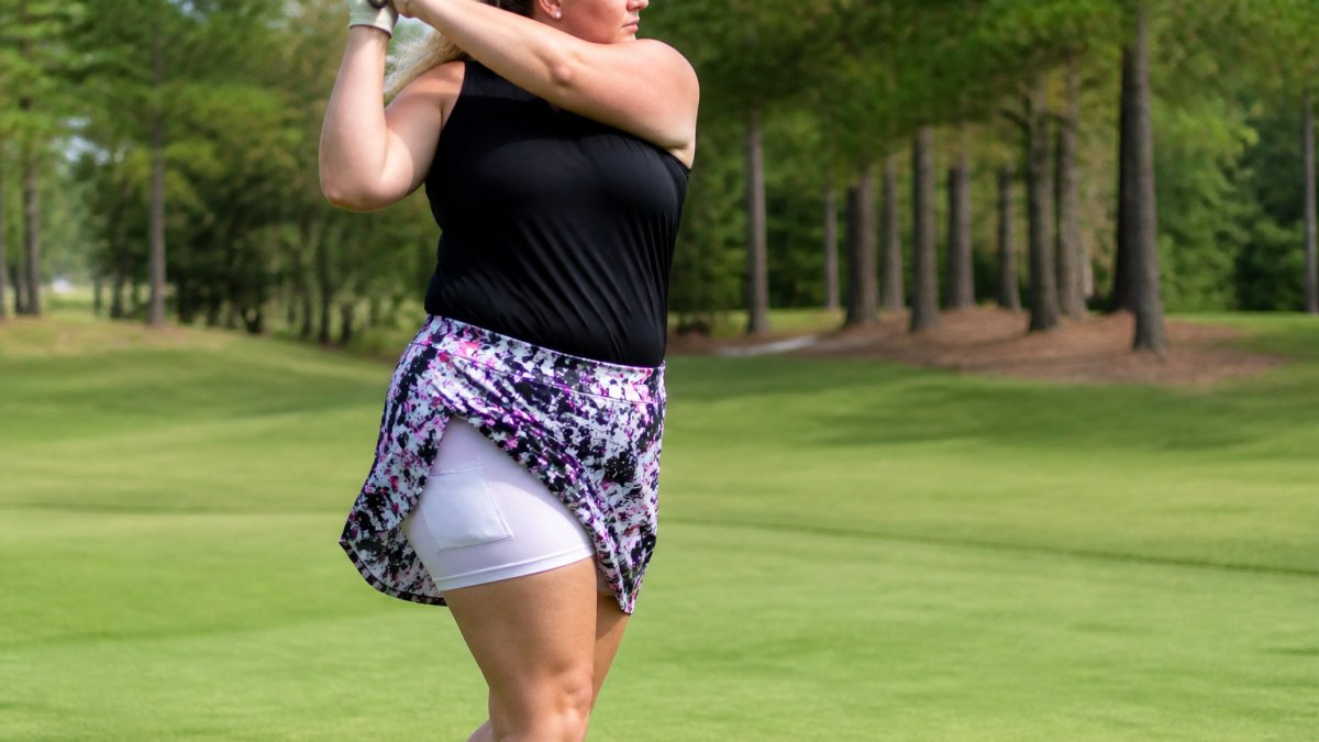 Woman golfer in plus-size skort mid-swing on golf course, showing movement and freedom in fabric with visible pockets and built-in shorts underneath, natural daylight