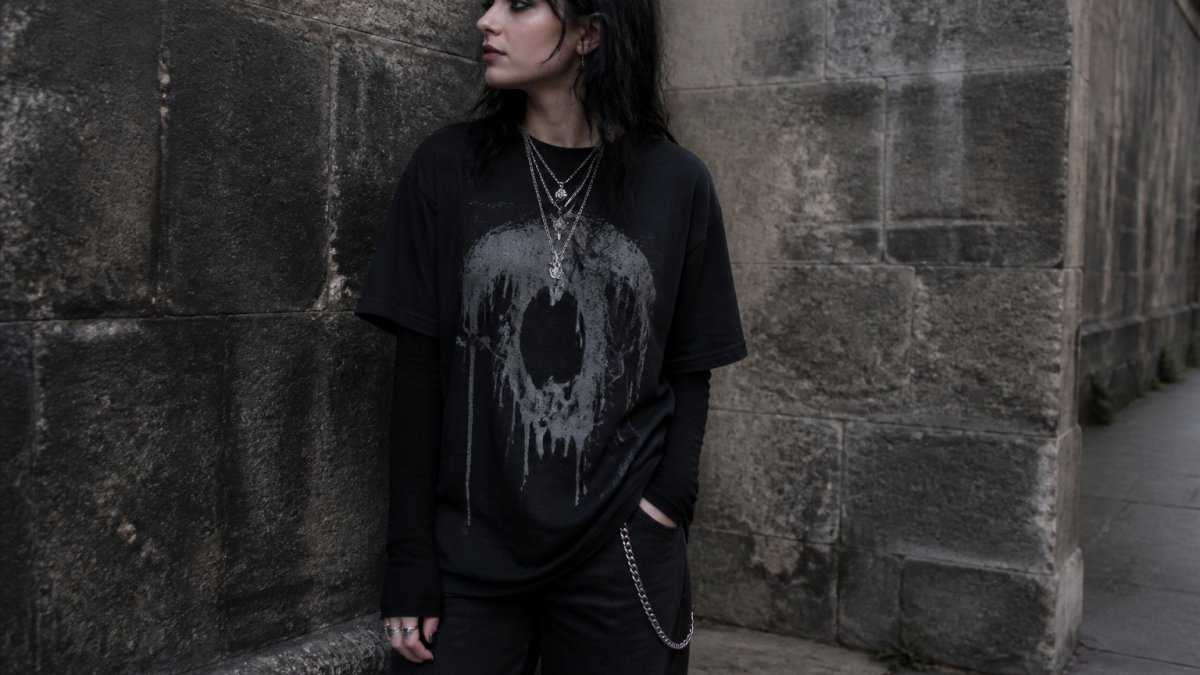 Person wearing a layered black graphic tee and silver chains against a moody urban stone wall backdrop