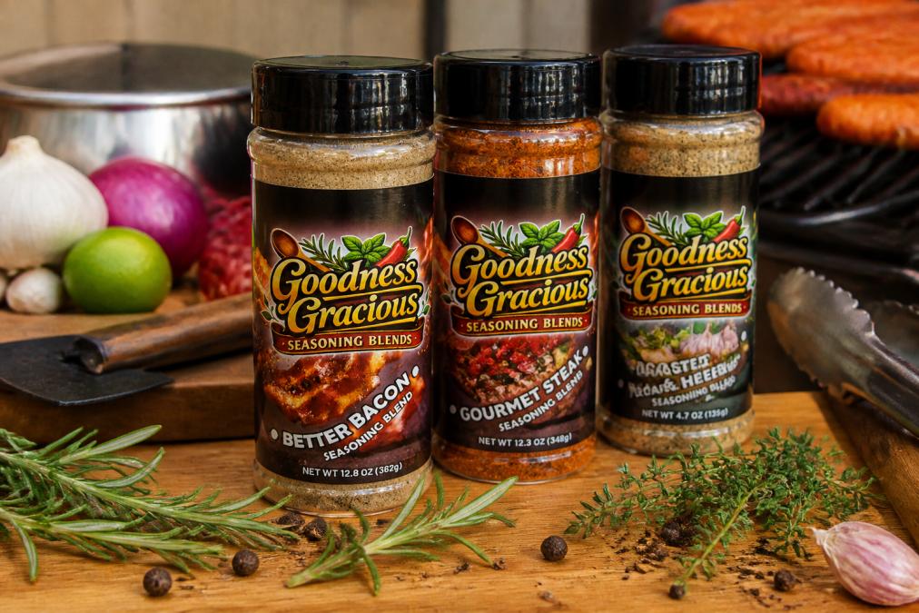 Close-up of three glass jars filled with different colored spice blends on a wooden kitchen counter with fresh herbs, peppercorns, and grilling tools arranged nearby