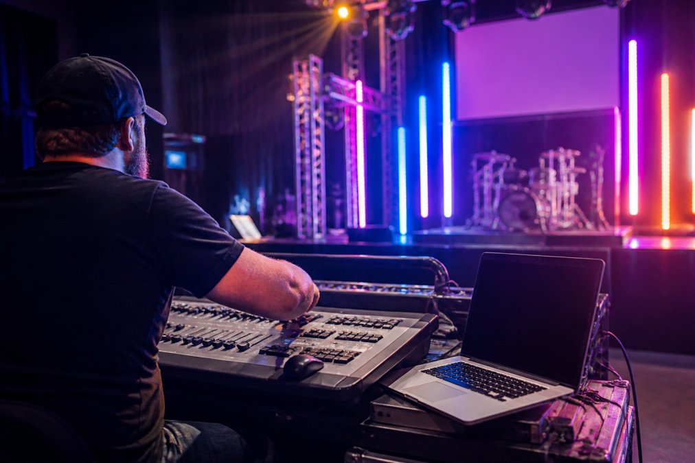 A lighting technician adjusting faders on a professional console in a dimly lit venue, a laptop running DMX software visible beside them, controlling colorful smart light bars mounted on stage trusses