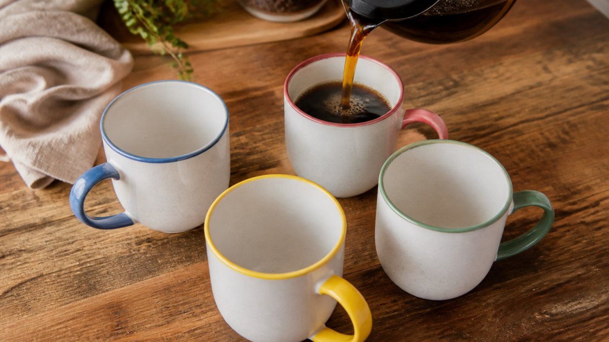 Overhead view of four colorful ceramic mugs on a wooden table while coffee is poured into one during a kitchen scene