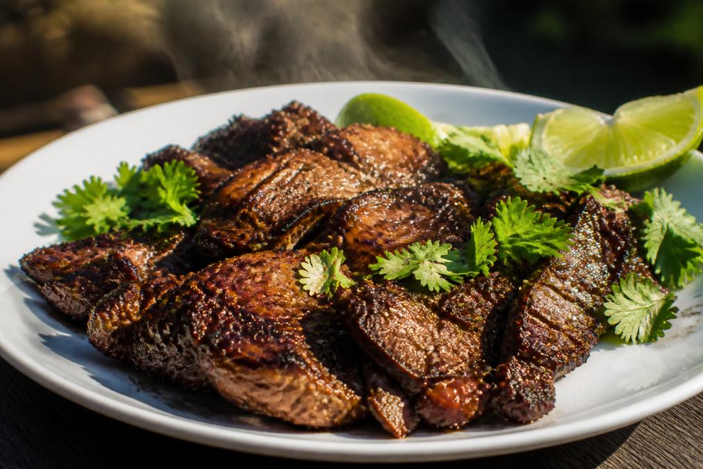 Close-up of freshly grilled meat with deep caramelized crust and grill marks, glistening with marinade, steam rising from the surface, served on a white plate with fresh cilantro garnish and lime wedges