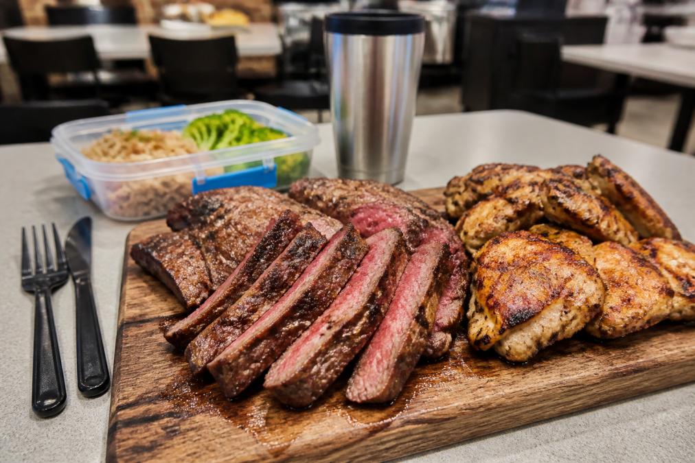 Close-up of sliced grilled flank steak and chicken thighs arranged on a wooden board with visible grill marks, next to a lunch container and utensils, in a bright office breakroom