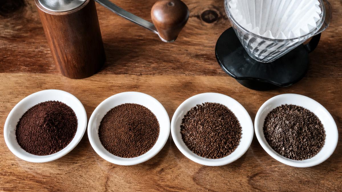 Overhead shot of four small white saucers on a wooden counter, each holding a different coffee grind texture from fine to coarse, with a hand grinder and a paper filter cone beside them