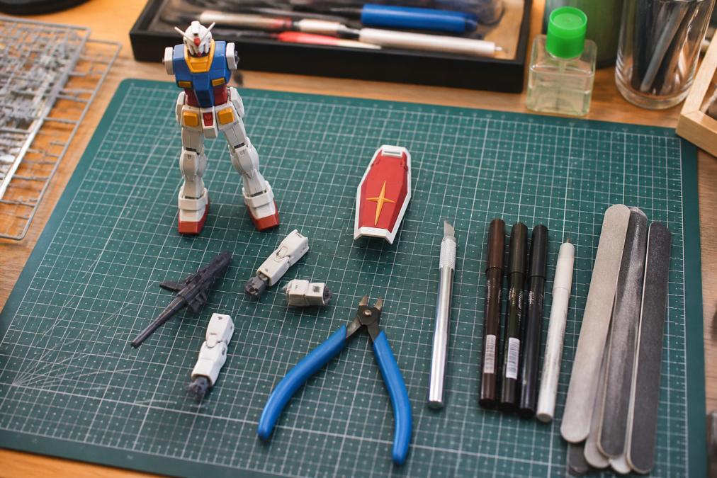Overhead shot of a Gunpla hobby workspace with cutting mat, nippers, hobby knife, sanding sticks, panel line markers, and a partially assembled Gundam model arranged neatly on a wooden desk