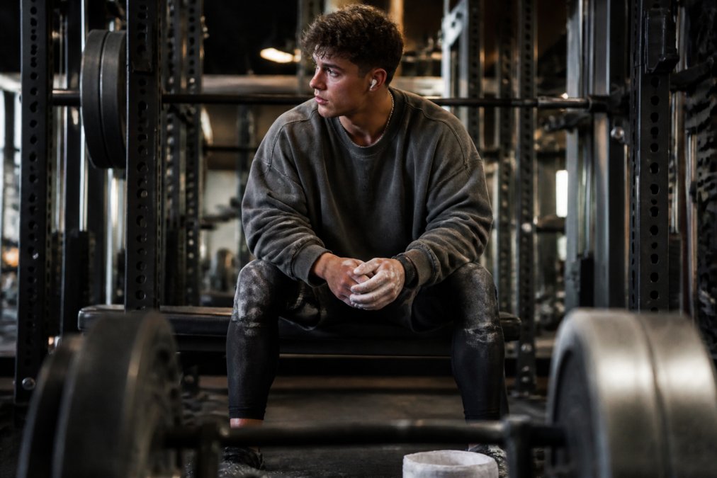 A weightlifter in an oversized snow-washed pump cover resting on a bench between heavy squat sets, gym chalk and a loaded barbell visible in the foreground, warm overhead lighting