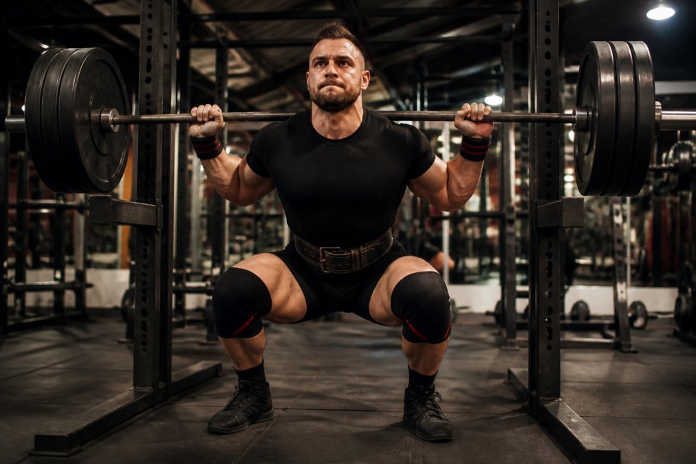 Powerlifter wearing wrist wraps, knee sleeves, and lifting belt during heavy squat, demonstrating proper joint support and ergonomic positioning with engaged core and neutral spine in well-lit commercial gym
