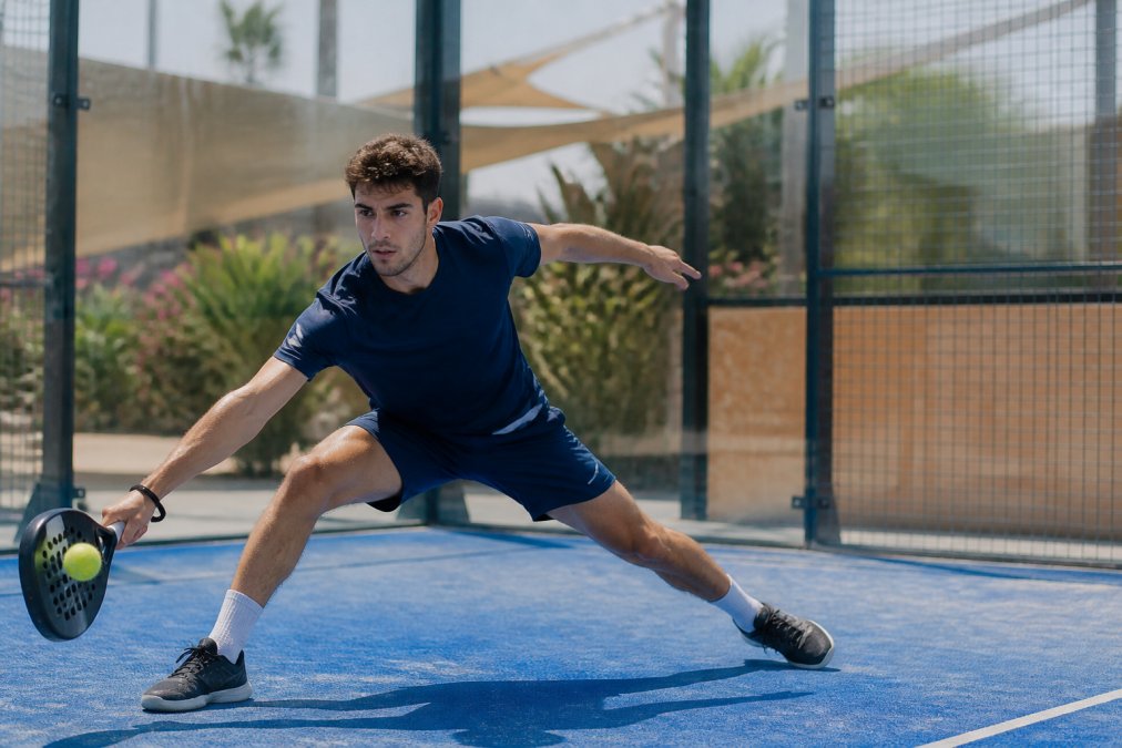 Professional padel player executing a lateral movement shot on outdoor court, wearing technical padel clothing with visible four-way stretch and moisture-wicking fabric, natural outdoor lighting showing athletic motion
