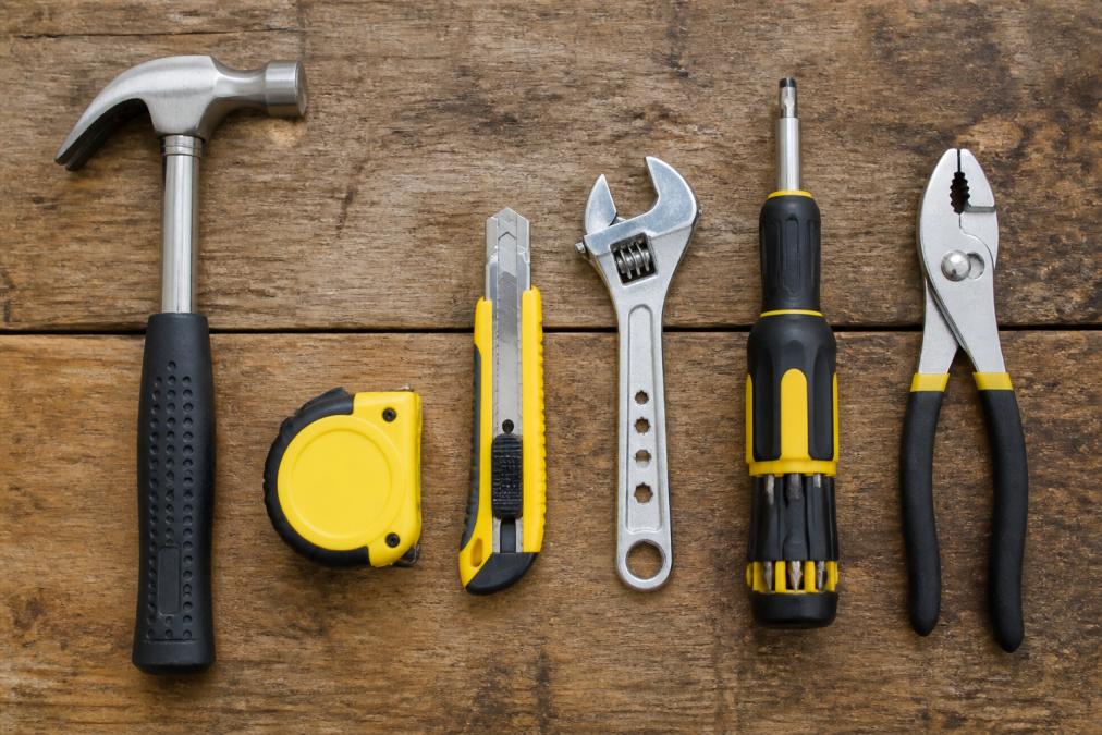 Flat lay of common hand tools arranged neatly on a wooden workbench including a claw hammer, tape measure, utility knife, adjustable wrench, multi-bit screwdriver, and slip-joint pliers, all well-lit against a natural wood surface